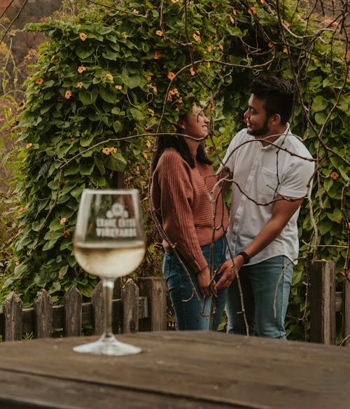 Free Side view content ethnic couple in casual clothes bonding and looking at each other while standing in lush park near table with glass of white wine Stock Photo