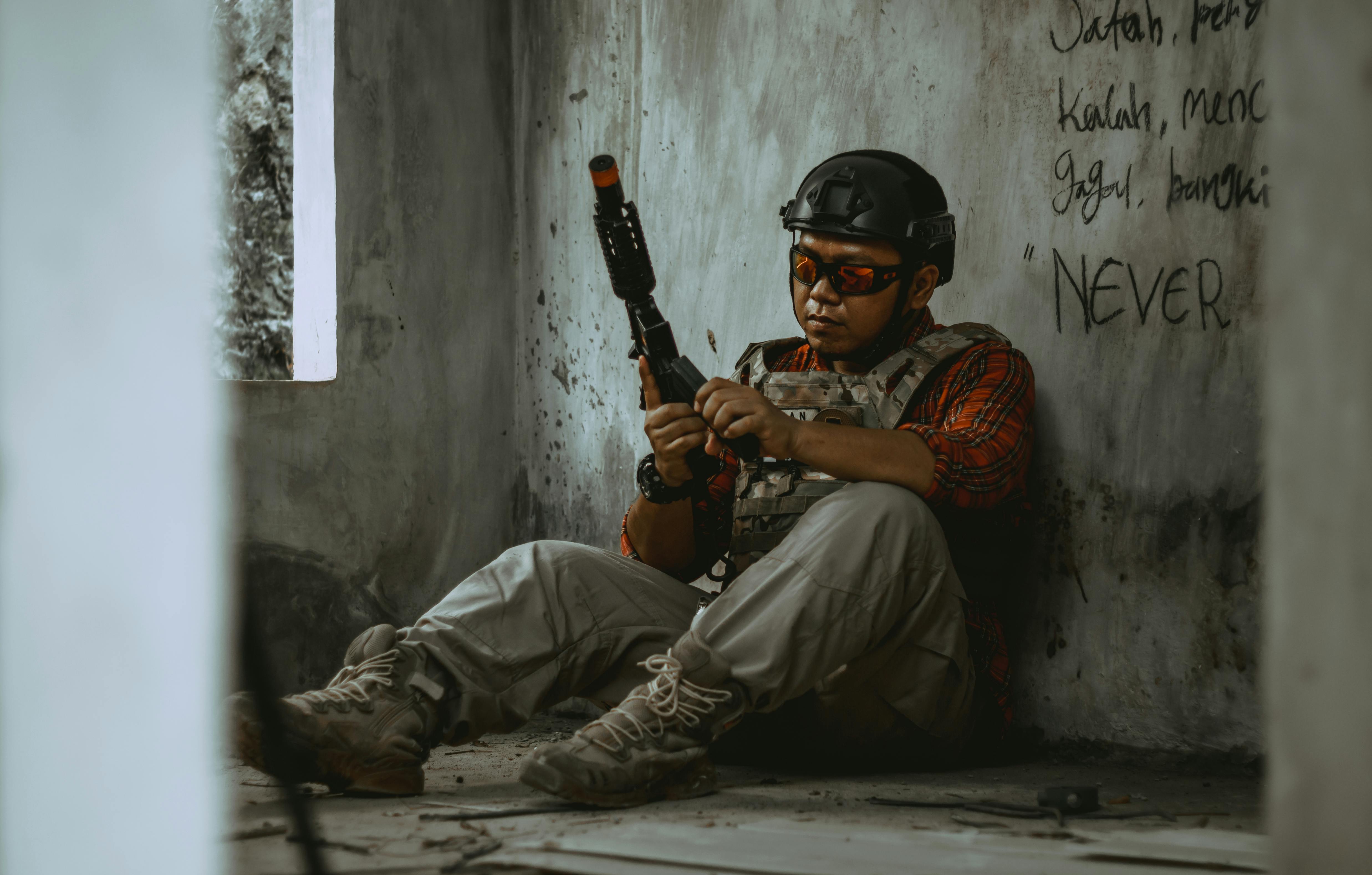 A soldier in tactical gear rests indoors, adjusting his weapon. Dim lighting sets a tense mood.