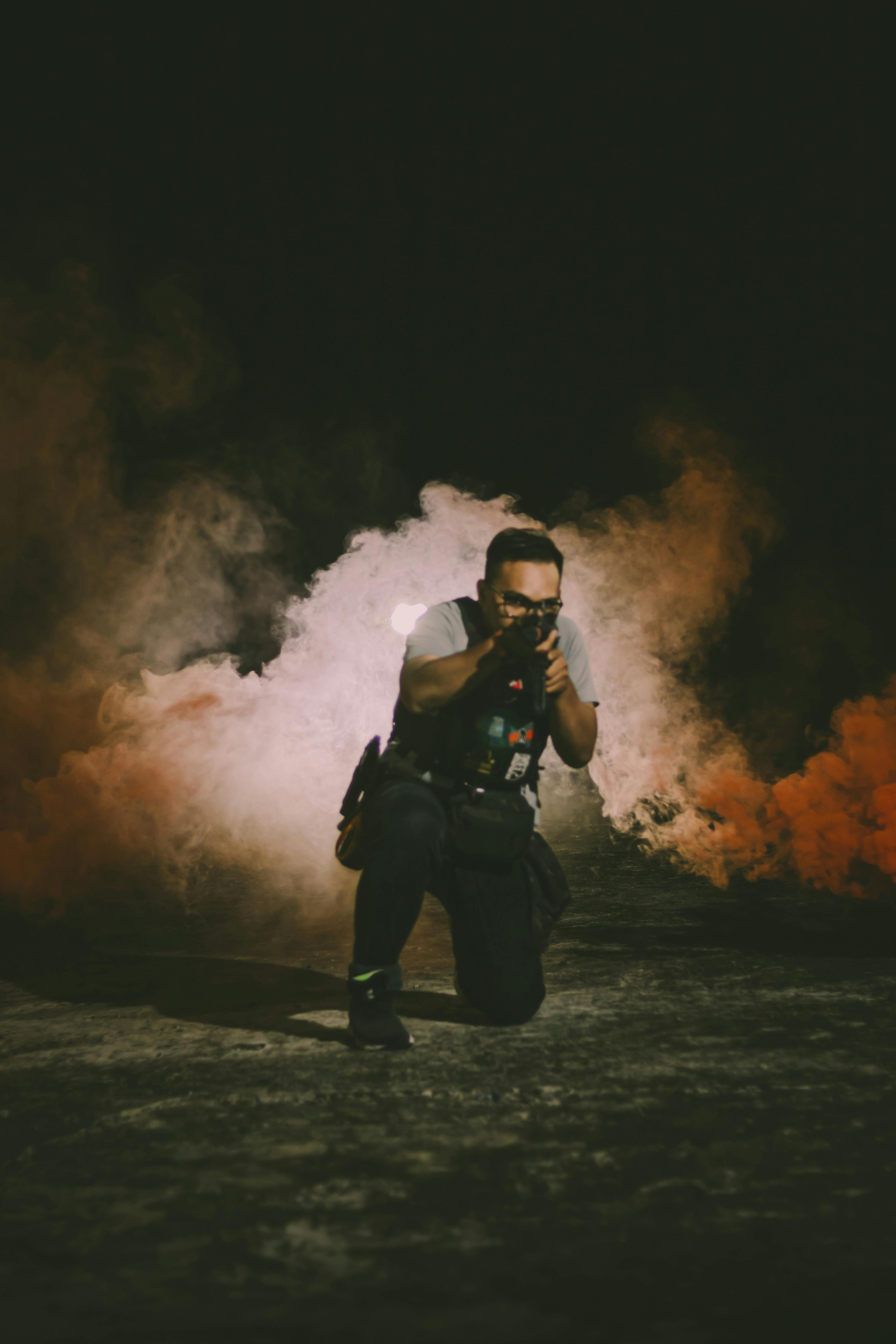 Man crouching in smoke with tactical gear and weapon aiming outdoors.