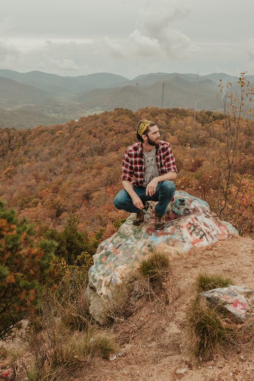 Full body of young bearded male traveler in checkered shirt and jeans sitting on haunches on rocky boulder and admiring picturesque nature in mountainous valley covered with lush autumn trees