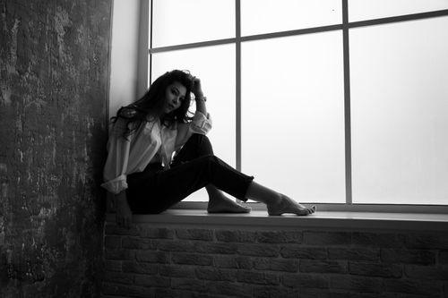 Free Black and white full body side view of pensive young barefoot female sitting on windowsill and looking at camera Stock Photo
