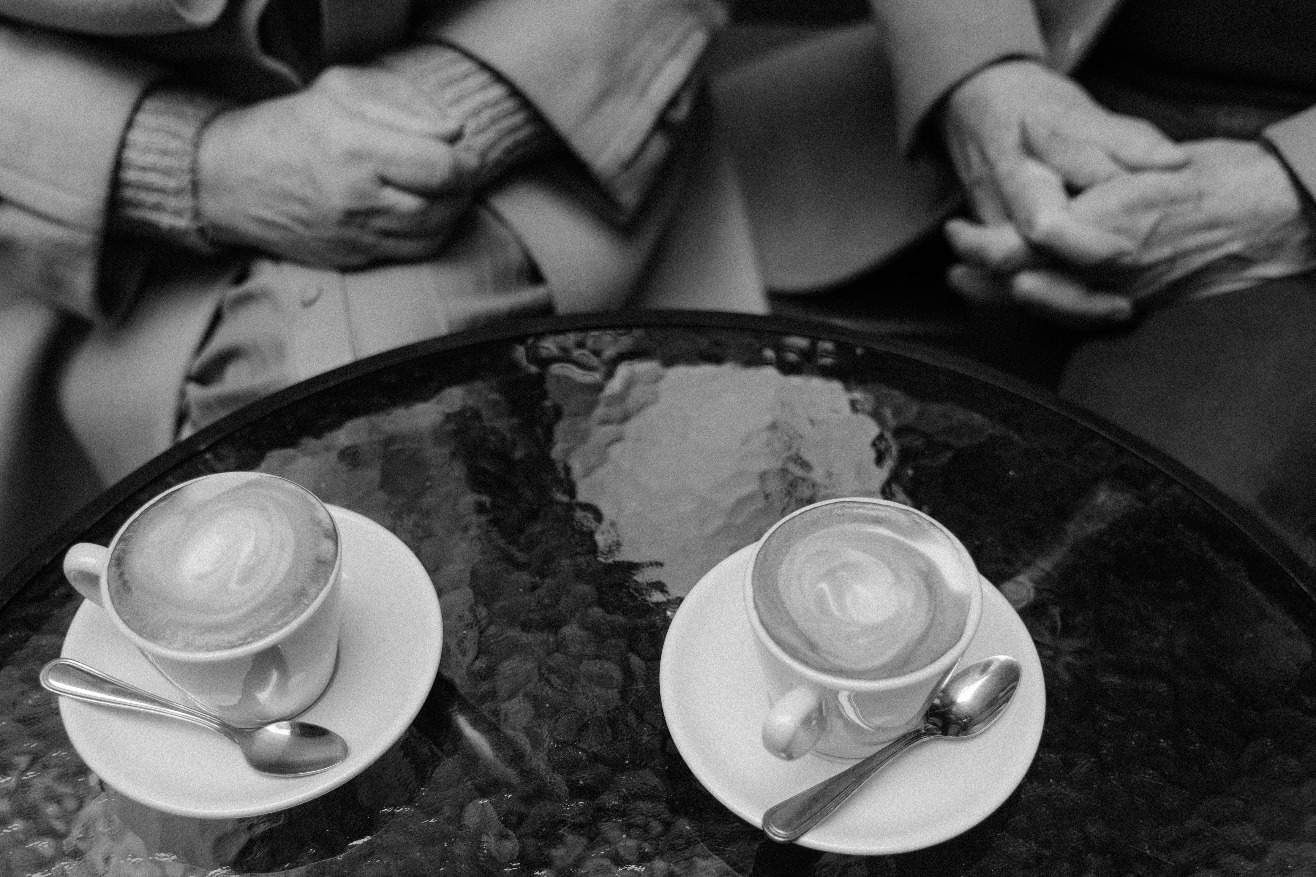 桌子上放着两张卡布奇诺咖啡的单色照片，背景里是两个人的手