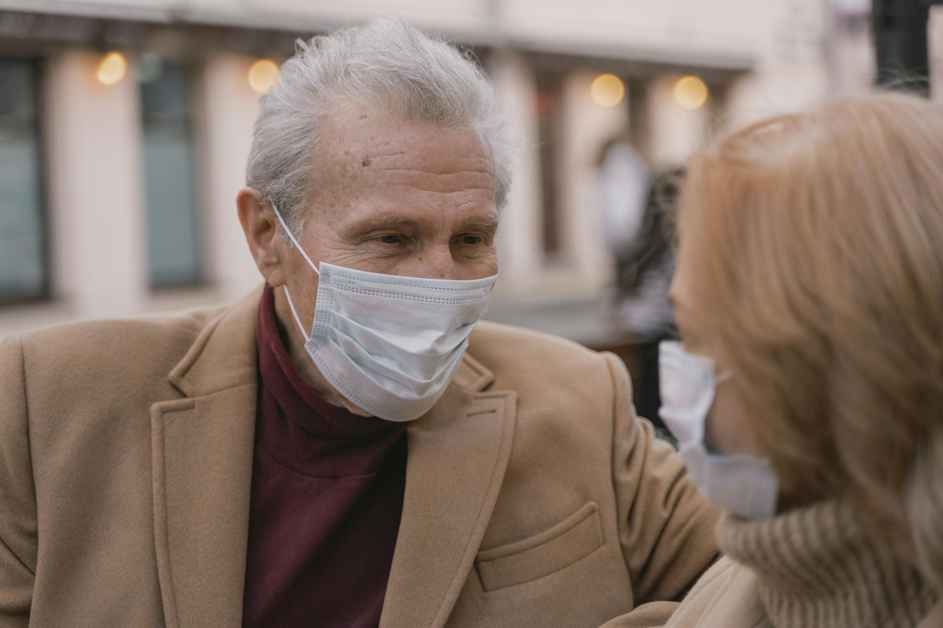 Elderly couple wearing masks and brown coats, sharing a moment outdoors in a safe and caring manner.