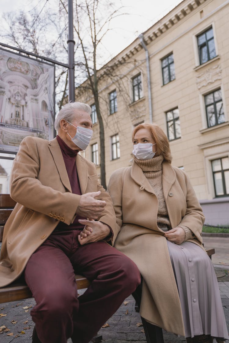 An Elderly Couple Sitting On A Bench With Facemasks On