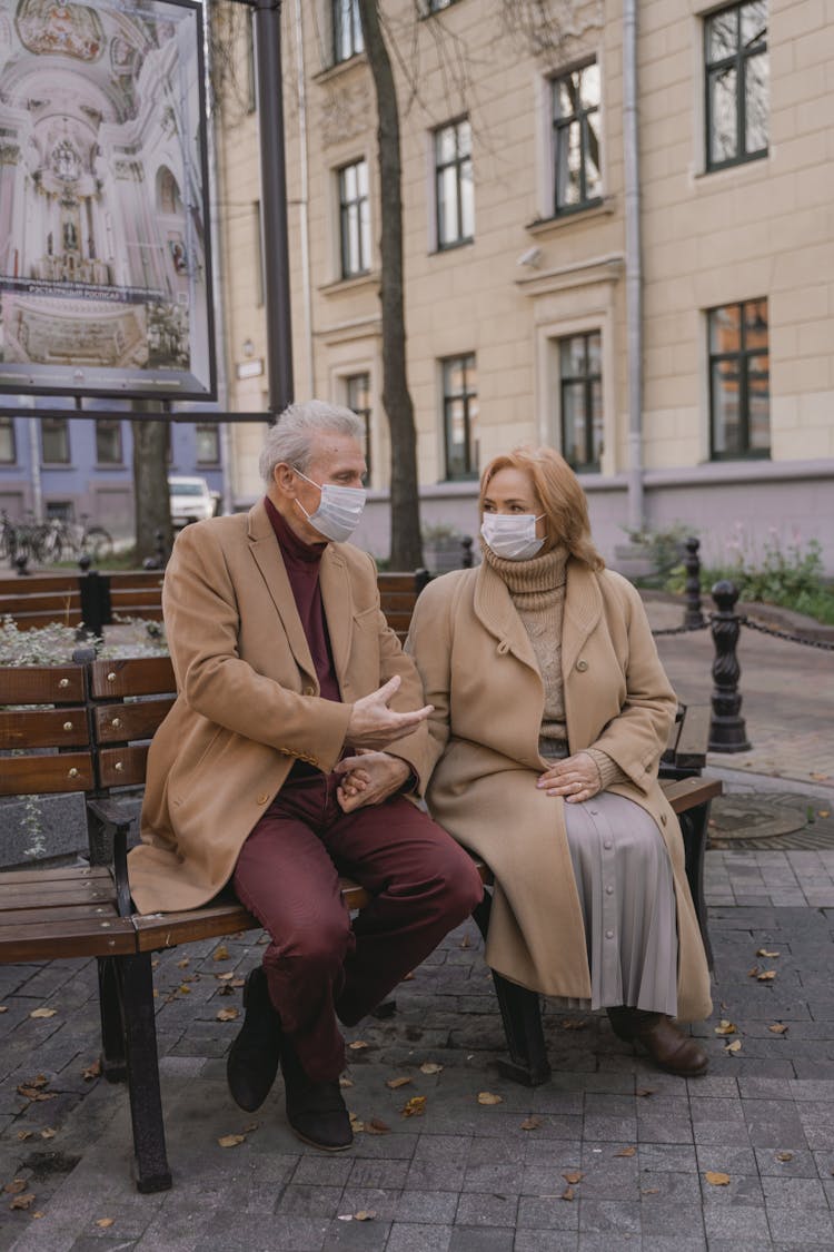 An Elderly Couple Sitting On A Bench Wearing Facemasks