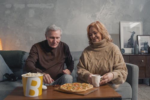 Smiling Elderly Couple Sitting by Table with Pizza and Popcorn