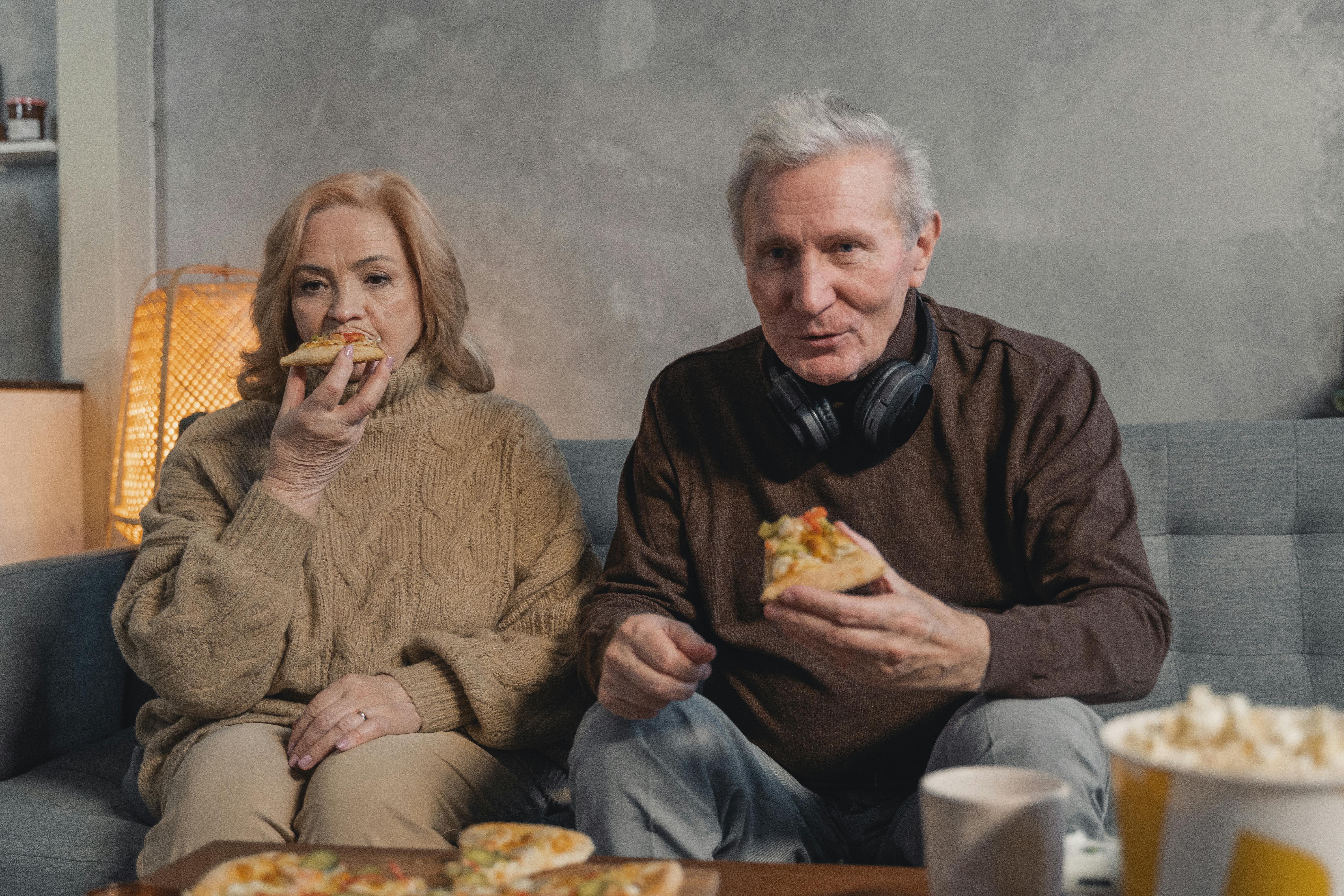 elderly couple sitting on couch eating pizza