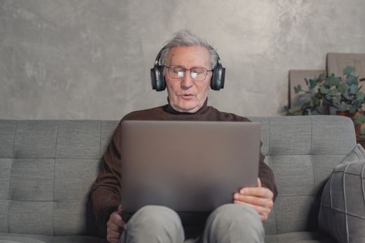 Senior man with headphones working on laptop at home, sitting on a gray couch.