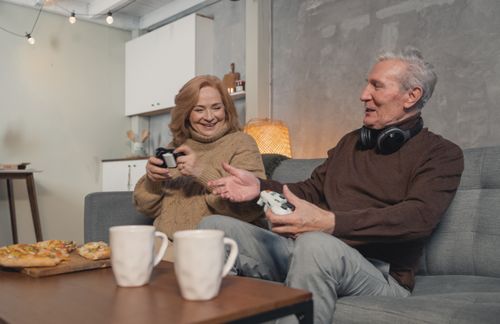 Free A joyful elderly couple playing video games with pizza and mugs nearby. Stock Photo