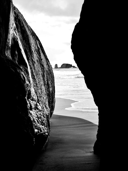 Black and white of rough rocks placed on sandy coast near wavy sea in daytime
