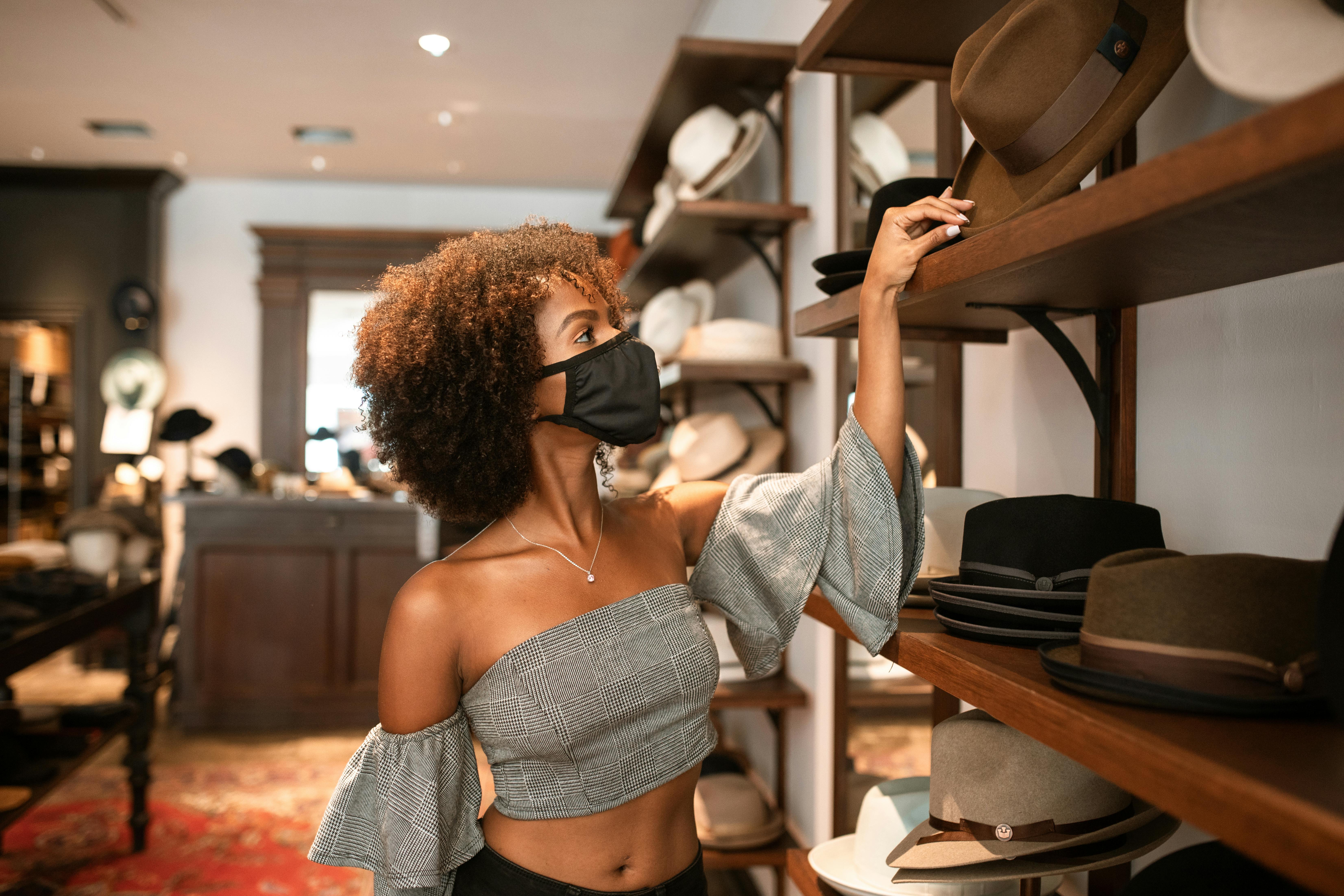 African American woman in mask chooses a stylish hat in a boutique store.