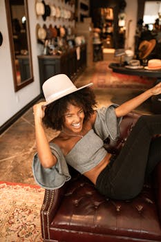 African American woman sitting happily in a stylish boutique, trying on hats and having fun.