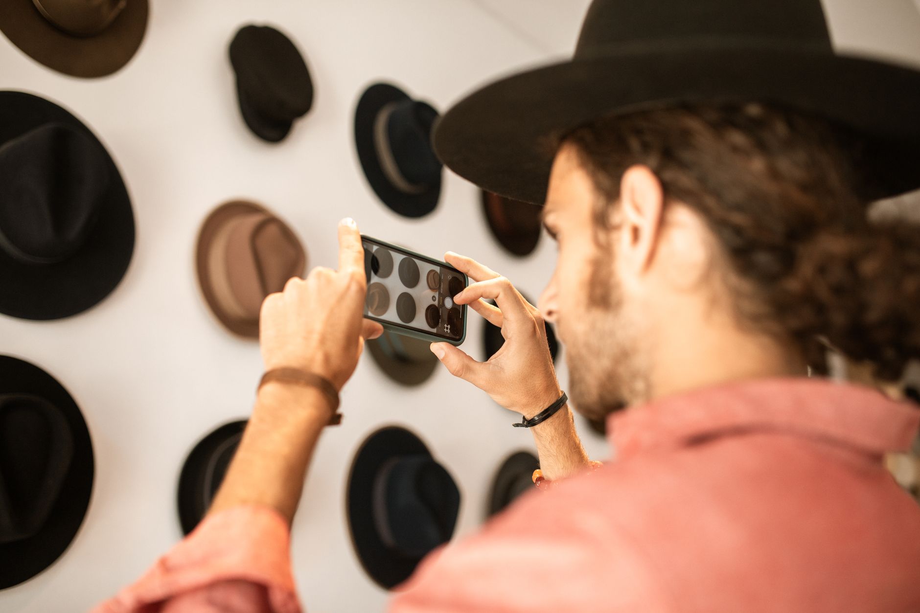 A man in a hat takes photos of a hat display with a smartphone indoors.