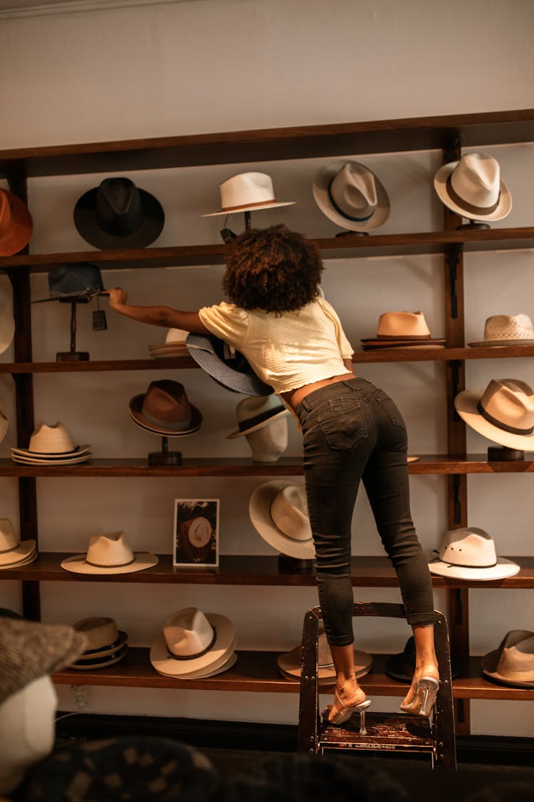 A Woman Standing On A Ladder While Reaching A Hat On A Wooden Shelves