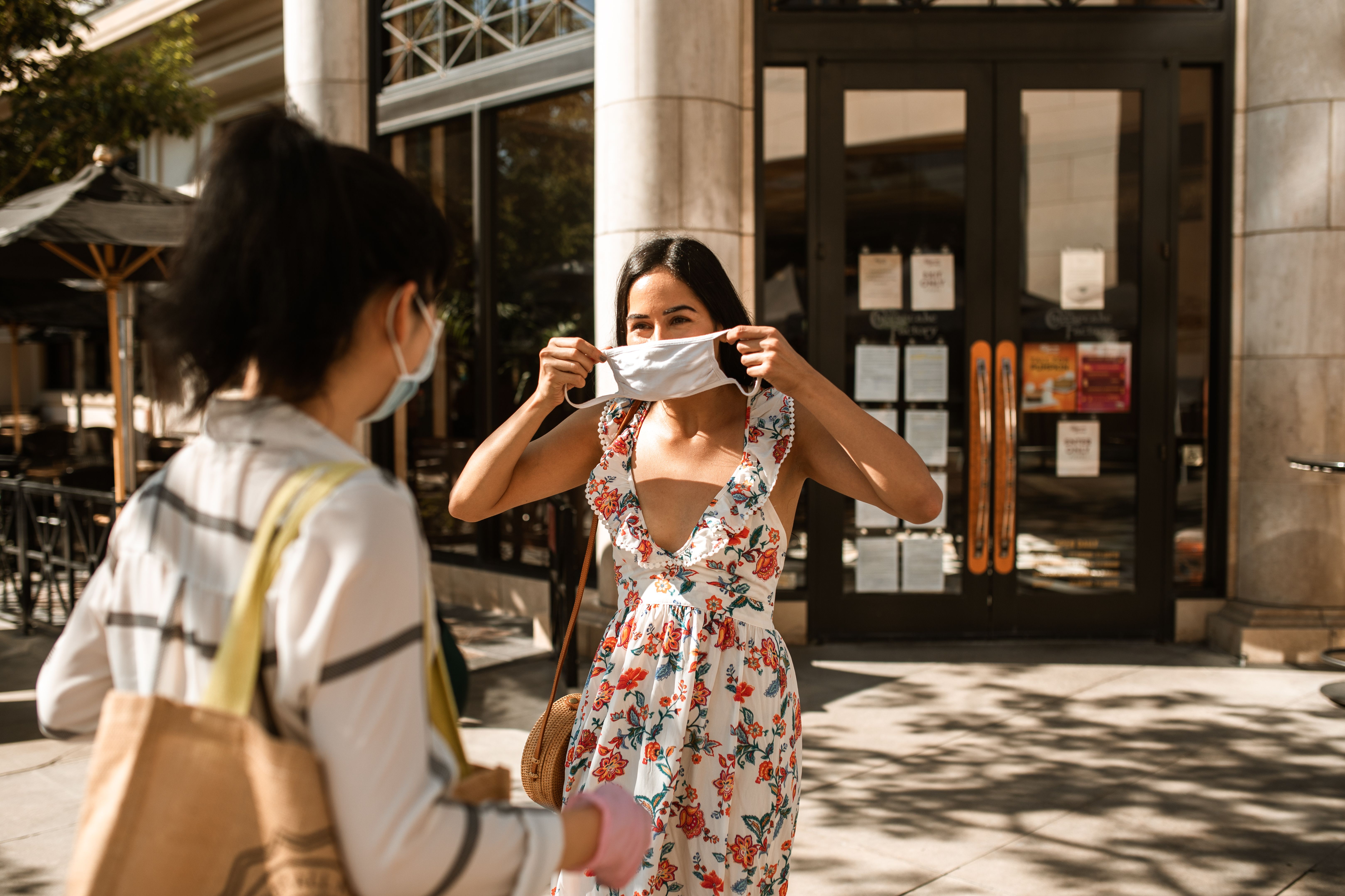 Two women in floral dresses meet outdoors, adjusting face masks during a sunny day.