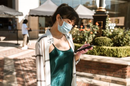 Free Young Asian woman wearing a mask, using a smartphone outside, reflecting urban life during the pandemic. Stock Photo