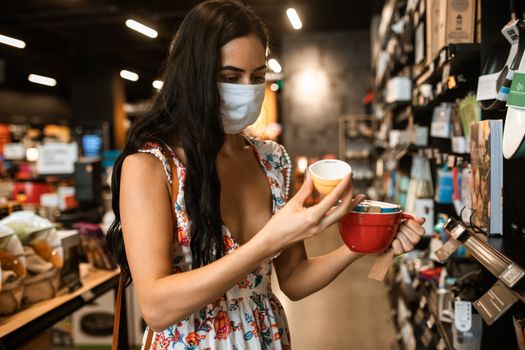 一位穿着花裙、戴着口罩的女子在室内购物，正考虑购买陶瓷杯