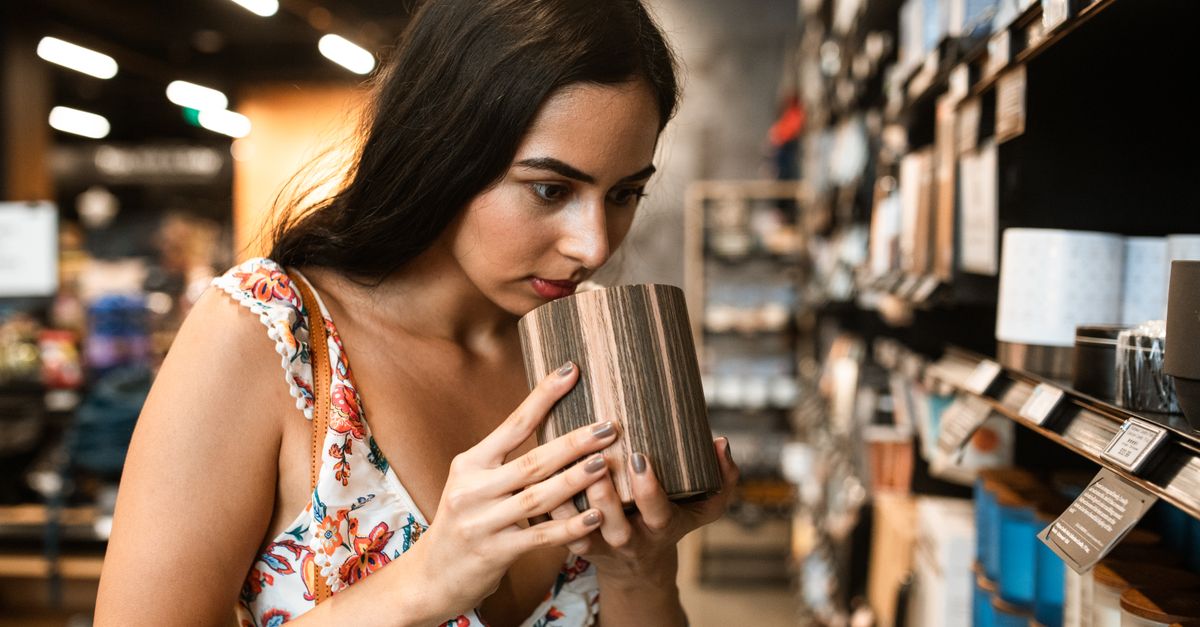 Young woman in floral dress smelling a candle in a cozy store setting, engaging in shopping.