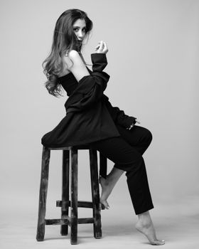 Stylish black and white portrait of a barefoot woman in a black suit posing on a stool in a studio.