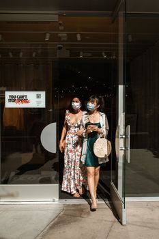 两名穿着时髦的女性戴着口罩走出商店，这代表着大流行时代的购物