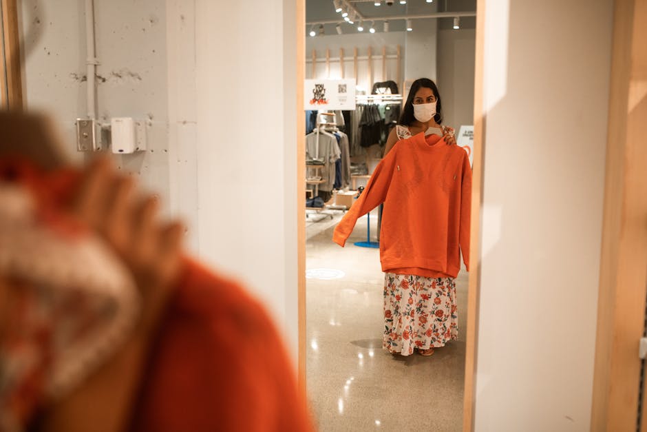 A woman wearing a mask checks her reflection while holding a sweatshirt in a stylish store.