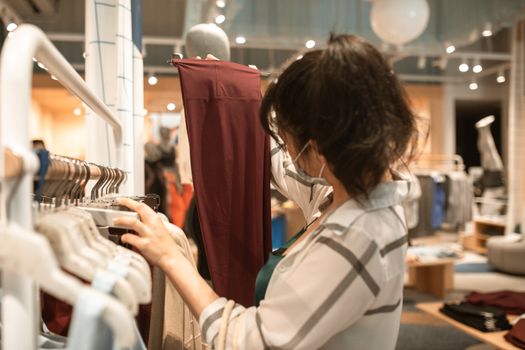 一位戴口罩的女子在商店里挑选衣服，最后选了一条酒红色的裤子