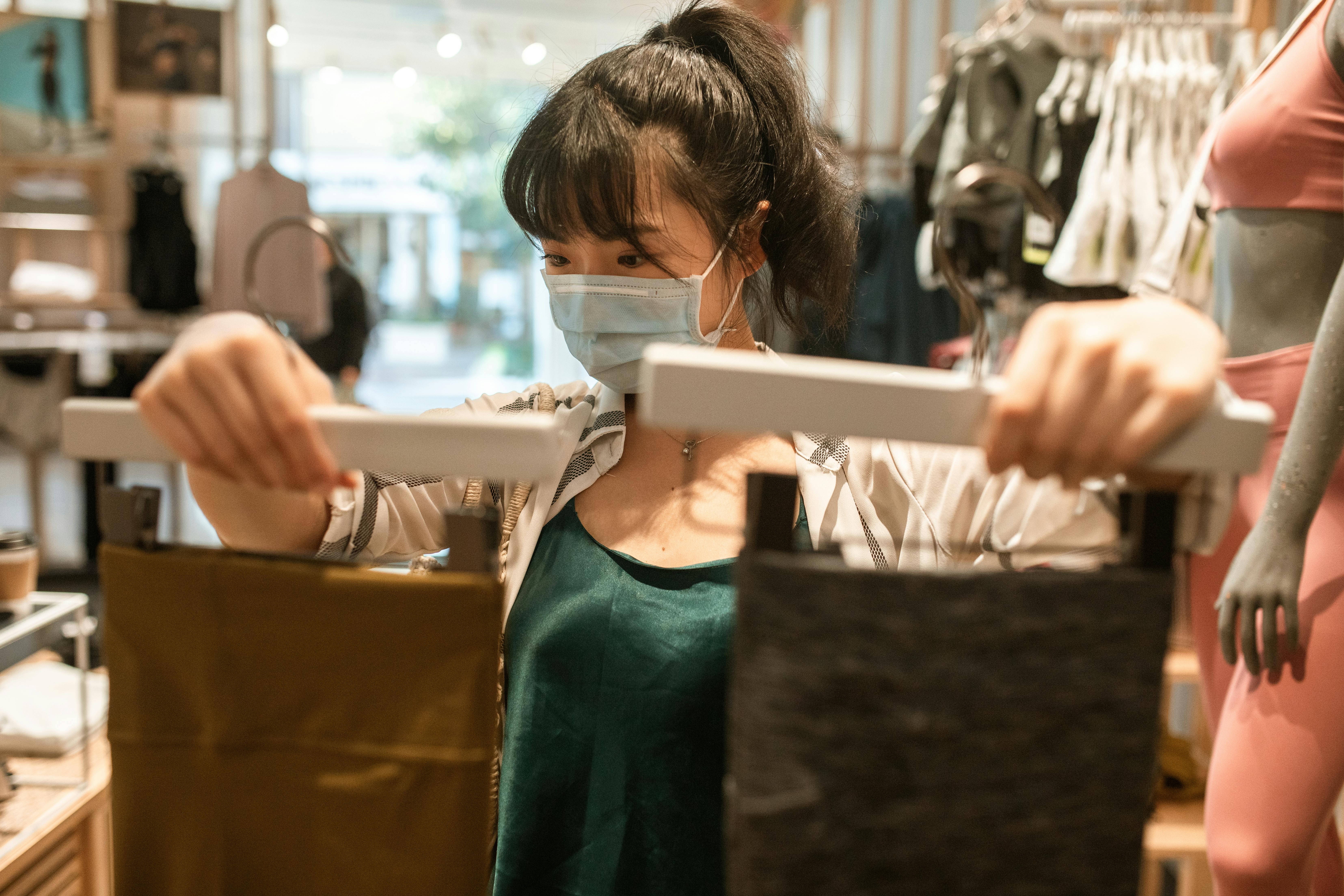 Masked woman choosing brown pants in clothing store, reflecting pandemic shopping norms.