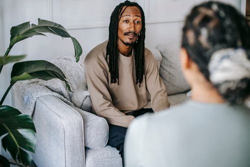 Free Content African American male with dreadlocks wearing casual outfit having conversation with female friend while sitting on couch in cozy living room Stock Photo