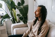 From above side view of pensive bearded African American male with dreadlocks sitting on sofa near green plant in cozy therapist office