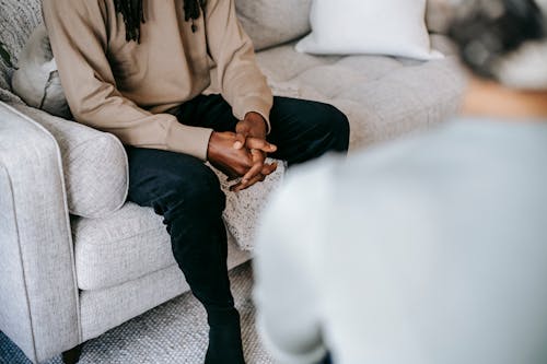 Unrecognizable ethnic male client with hands clasped sitting on sofa near blurred female therapist during psychotherapy appointment in cozy office