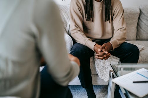 Unrecognizable African American male patient with clasped hands sitting on sofa near blurred anonymous therapist during visit in psychotherapy office