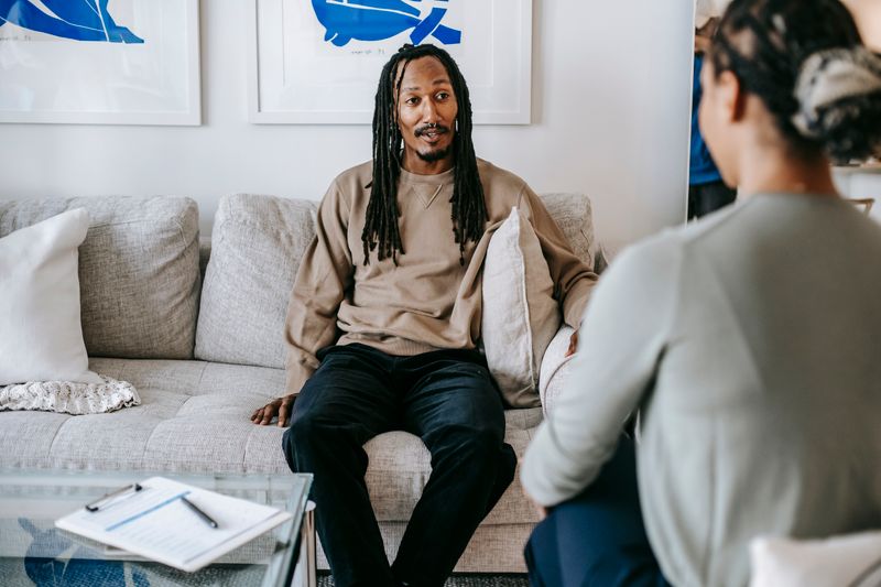 A young Black man with shoulder-length dreadlocks sits on a couch in a sunlit room in calm composed conversation with a female therapist; a clipboard rests on the glass coffee table between them.