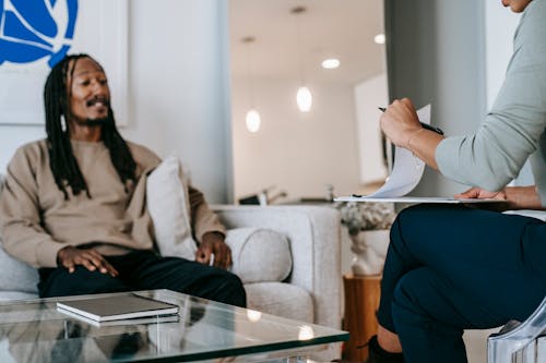 African American male with dreadlocks sitting on sofa in front of unrecognizable female therapist with clipboard during psychotherapy appointment in office