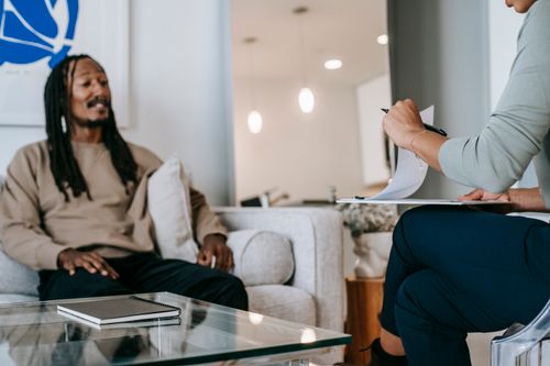 Free African American male with dreadlocks sitting on sofa in front of unrecognizable female therapist with clipboard during psychotherapy appointment in office Stock Photo