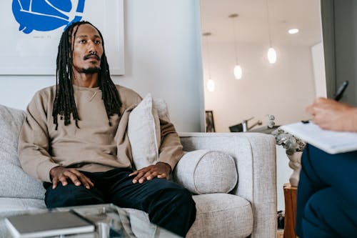 Pensive African American male client with dreadlocks sitting on couch with hands on laps near crop psychologist with clipboard during appointment