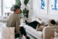 Ethnic female therapist with clipboard sitting near male client lying on couch with clasped hands during psychotherapy session in cozy office