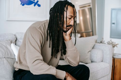 Free Side view of melancholy African American male with dreadlocks leaning on hand while sitting on couch during therapy session on blurred background Stock Photo