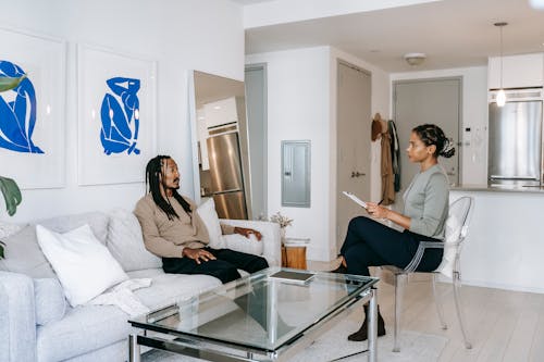 Concerned African American male in smart casual clothes sitting on comfortable sofa and sharing problems with empathetic female consultant in contemporary psychotherapy center