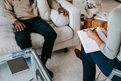 Free From above crop African American male in casual clothes sitting on comfy couch and answering professional female consultant questions in modern psychotherapy office Stock Photo