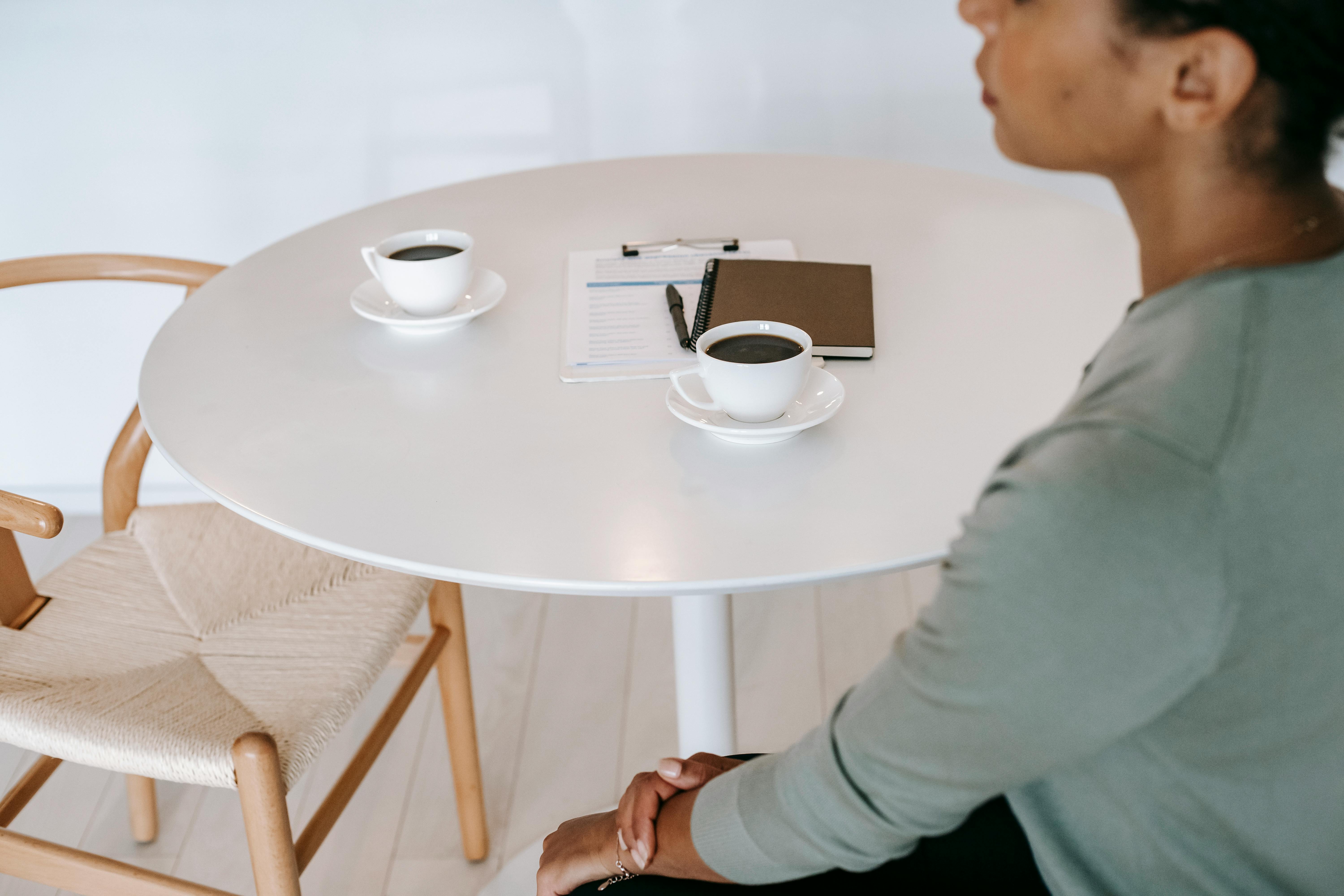 High angle crop ethnic female sitting at round table with cups of aromatic black coffee and diary in light room