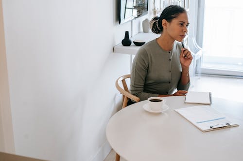 Concentrated ethnic female HR interviewer or psychologist in formal clothes sitting at round table with pen and notepad while looking away in contemplation
