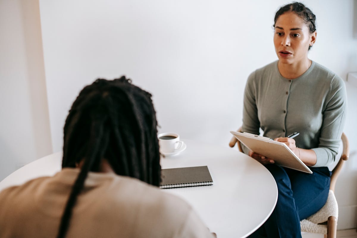 Focused ethnic female counselor wearing formal outfit sitting at table and discussing treatment method with male patient while taking notes in clipboard