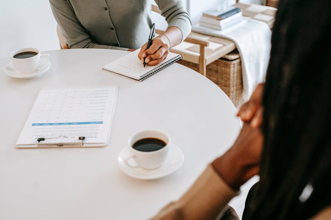 Free From above crop multiracial female interviewer or psychologist in formal wear asking questions and taking notes in planner while talking to black man and sitting together at table with coffee Stock Photo