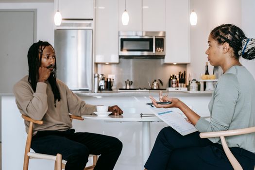 Professional ethnic female counselor giving advices and discussing mental problems with unhappy African American male client while sitting together in modern kitchen