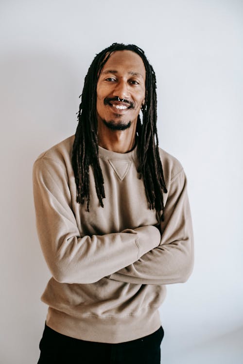Content adult African American male with dreadlocks wearing casual sweater standing with crossed arms against white wall and looking at camera with sincere smile