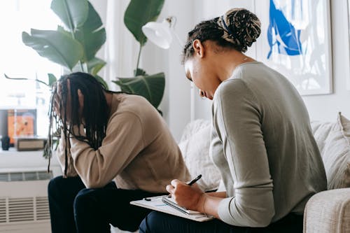 Professional female counselor taking notes in notepad while listening to depressed African American male client touching head in dismay in modern psychology center