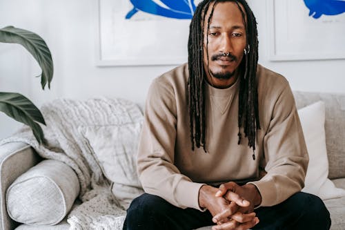 Free Crop thoughtful African American male in casual sweater with dreadlocks sitting with hands clasped on comfy couch in modern living room Stock Photo
