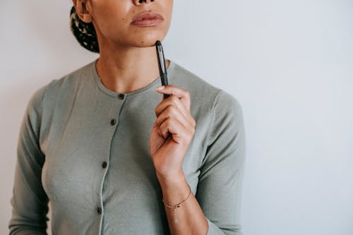 Crop anonymous ethnic female with dark hair in casual outfit touching face with pen and thoughtfully looking away against white wall