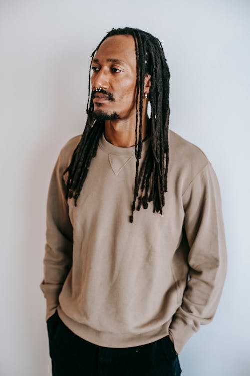Confident African American guy with dreadlocks and piercing in stylish clothes standing with hands in pockets and looking away against white background