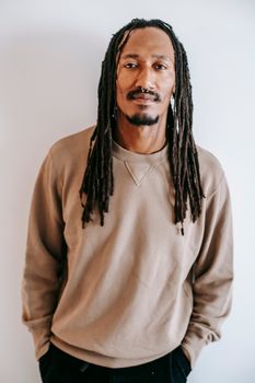 一位留着辫子的黑人男子身穿时尚服装，双手插在口袋里，站在白色背景前看着镜头